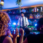 Young bartender preparing cocktails for cheering customers in a trendy bar with blue lights and hanging plants