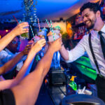 Happy friends raising cocktail glasses at nightclub bar counter, enjoying nightlife with bartender serving drinks