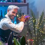 Professional bartender in a white shirt and black apron holds a steel shaker in his hands. Blurred bottles on shelves in background. High quality photo